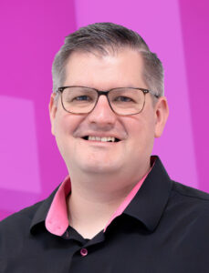 Eine Person mit kurzen Haaren und Brille lächelt in die Kamera. Sie trägt ein schwarzes Hemd mit rosa Kragen. Der Hintergrund ist in leuchtendem Pink mit geometrischen Mustern gehalten.