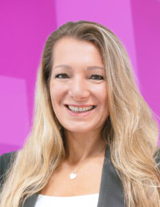 Lächelnde Frau mit langen blonden Haaren, trägt einen schwarzen Blazer und ein weißes Oberteil sowie eine herzförmige Halskette vor einem rosa Hintergrund.