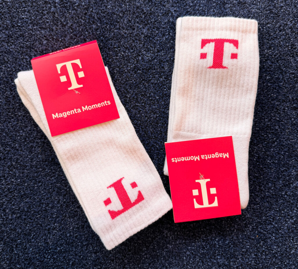 Auf einem dunkelblauen Teppich liegen zwei Paar weiße Socken mit rosa T-Mobile-Logos und roten Etiketten mit der Aufschrift „Magenta Moments“. Auf den Etiketten ist das T-Mobile-Logo in Weiß abgebildet.
